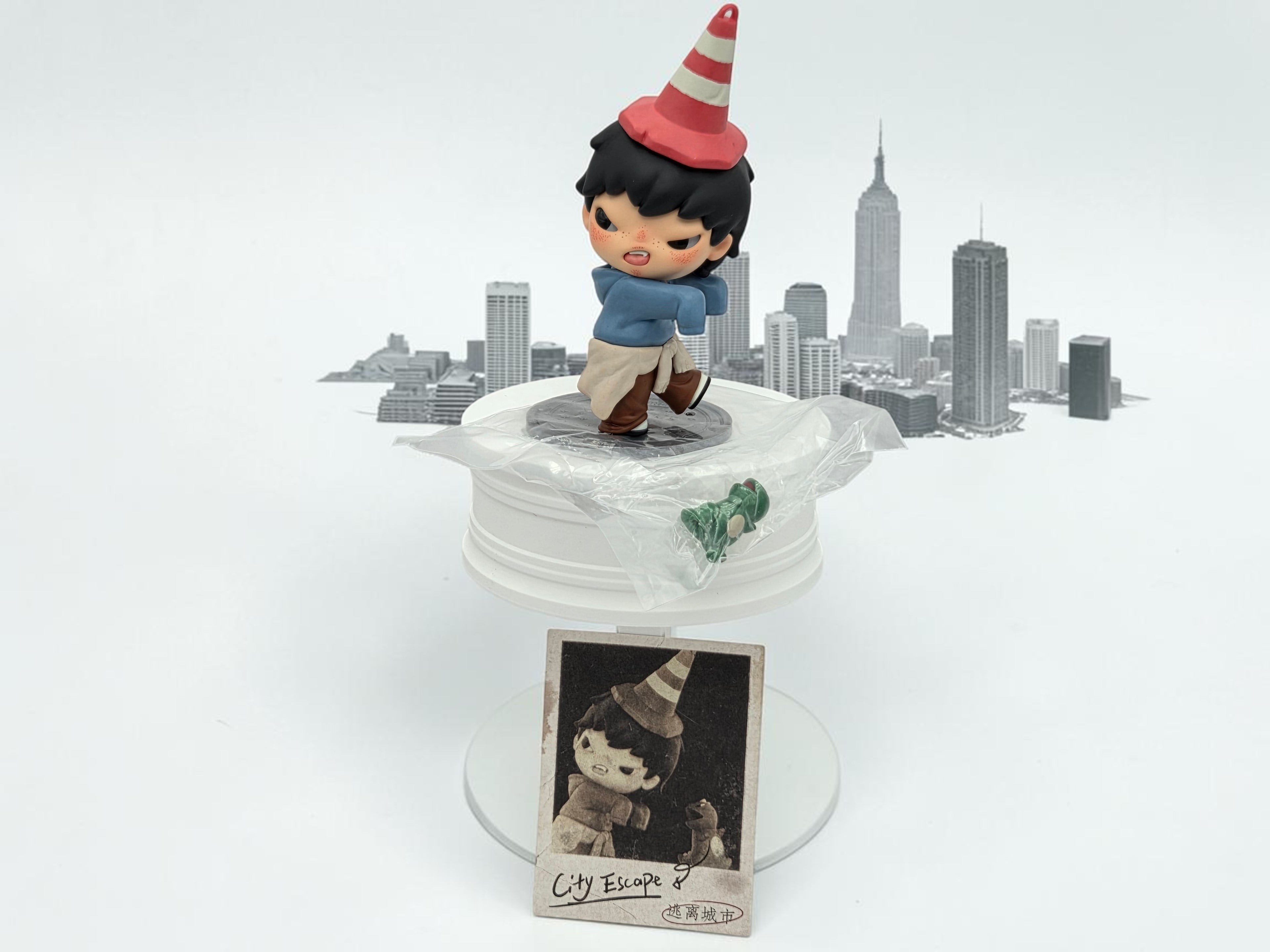 Momof2boyz Hirono Tamed Wildgrass figure with red and white striped cone hat, blue jacket, and tan pants, set against a cityscape backdrop.