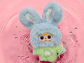Momof2boyz Shop Baby Three Petit Macaron bunny plush toy with blue hood, green body, and oversized expressive eyes.