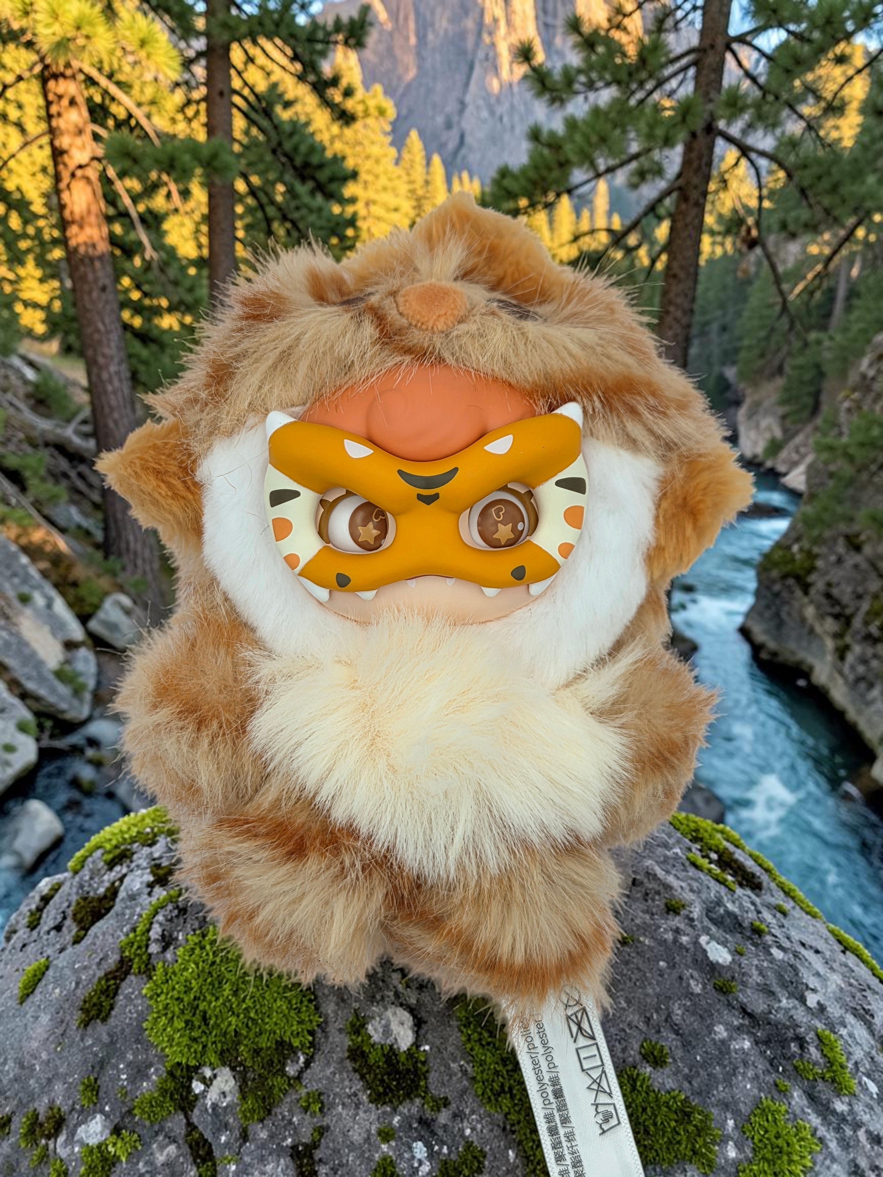 Momof2boyz Shop Mochi Little Monster Fortune Tiger plush keychain with removable mask on a mossy rock by a river.
