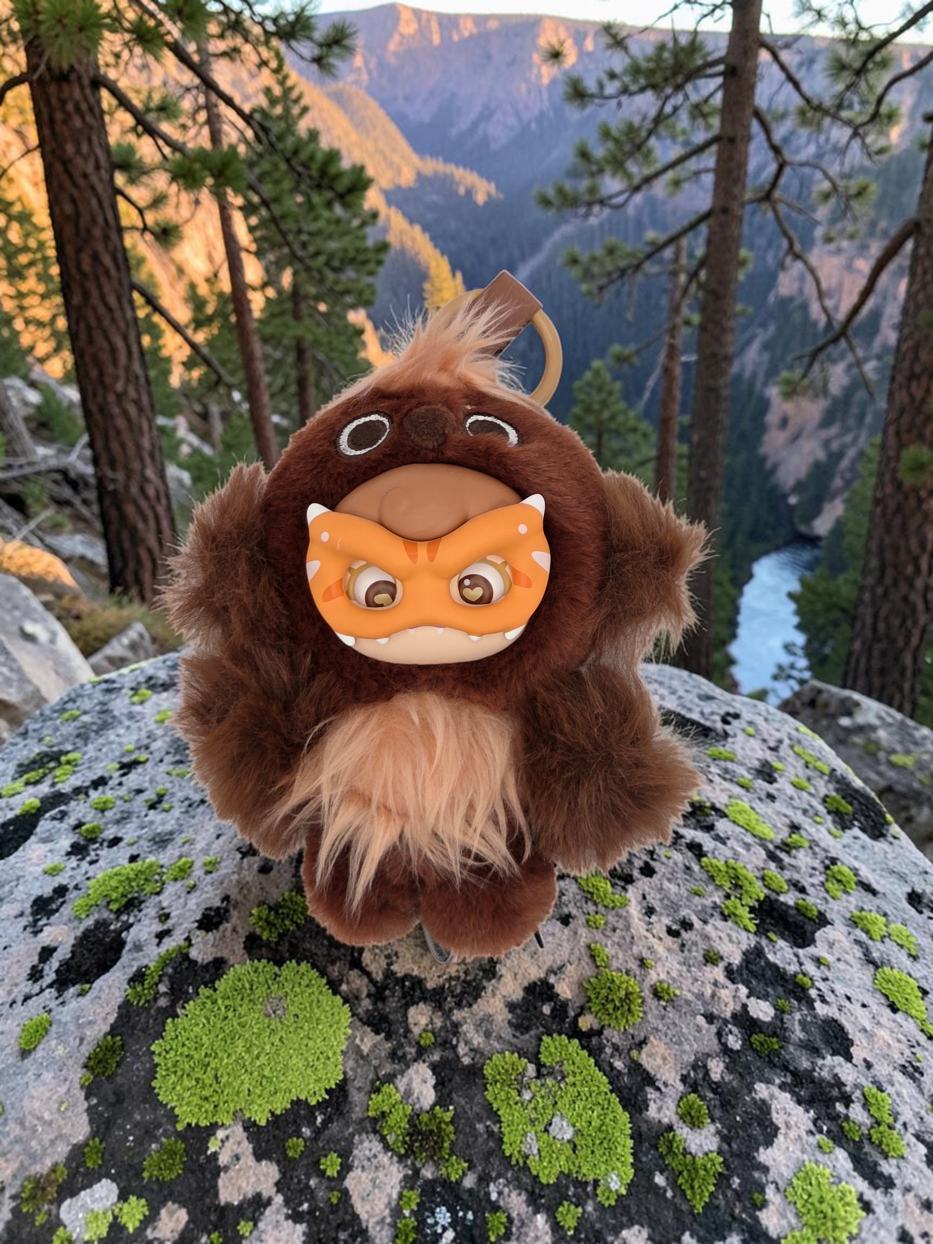 Momof2boyz Shop Mochi Little Monster plush keychain with a brown furry costume and orange tiger mask on a mossy rock
