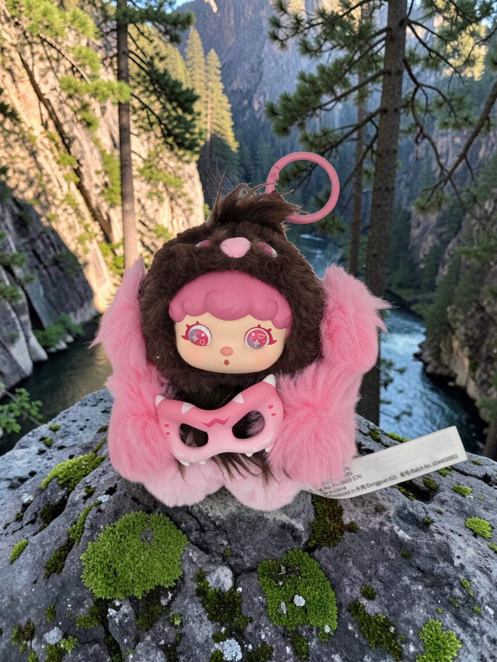 Momof2boyz Shop Mochi Little Monster plush keychain with pink hair and brown hood, posed on a mossy rock in nature.