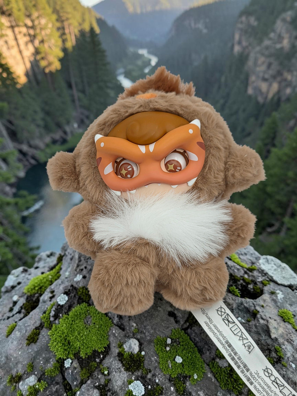 Momof2boyz Shop Mochi Little Monster plush keychain with brown fur and a removable orange monster mask on a cliff.