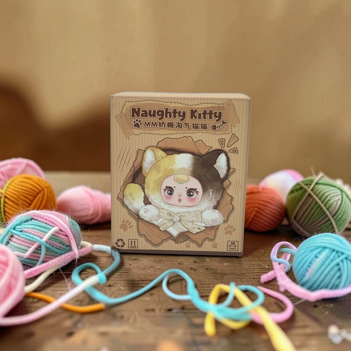 Momof2boyz Shop Naughty Kitty blind box plush toy in cardboard packaging surrounded by colorful yarn balls