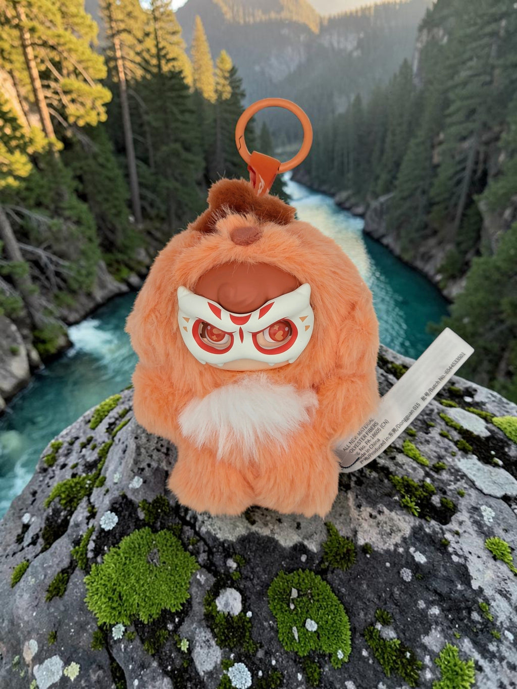 Momof2boyz Shop Mochi Little Monster plush keychain with orange fur and a decorative mask on a rocky riverbank.